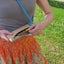 Frau, die eine hellgraue Filztasche mit einem blauen verstellbaren Riemen hält und auf einer Rasenfläche im Freien sitzt.